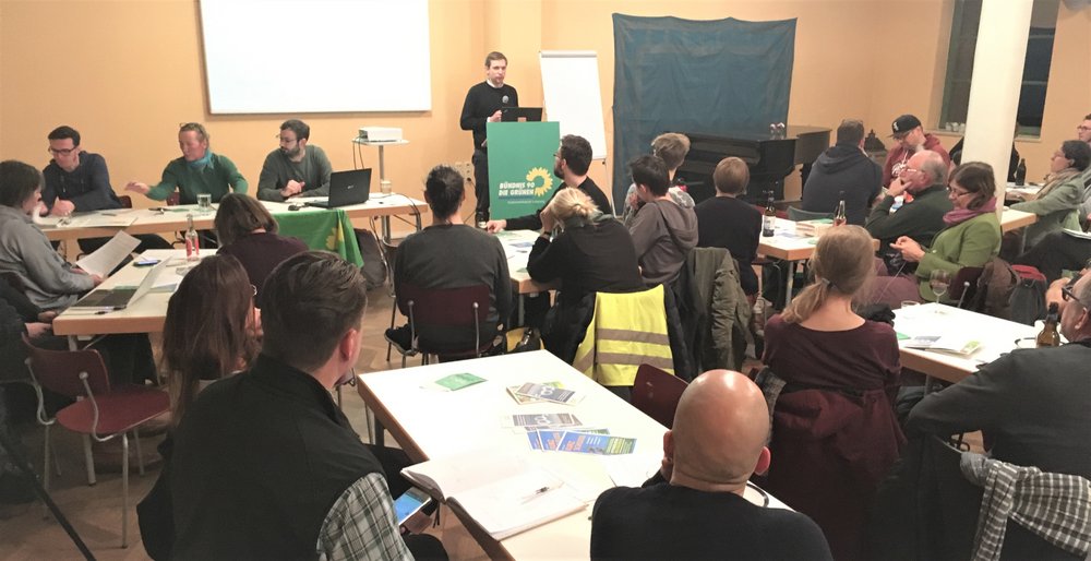 Auf dem Foto sieht man im hintergrund drei Personen neben einander am Tisch sitzen. Dahinter wirft ein Beamer ein Bild an die Wand. Daneben steht ein Mann am Rednerpult. Vor diesen sitzen ihnen zugewandt viele Zuschauer mit dem Rücken zum Betrachter.