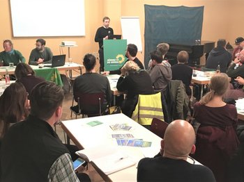 Auf dem Foto sieht man im hintergrund drei Personen neben einander am Tisch sitzen. Dahinter wirft ein Beamer ein Bild an die Wand. Daneben steht ein Mann am Rednerpult. Vor diesen sitzen ihnen zugewandt viele Zuschauer mit dem Rücken zum Betrachter.