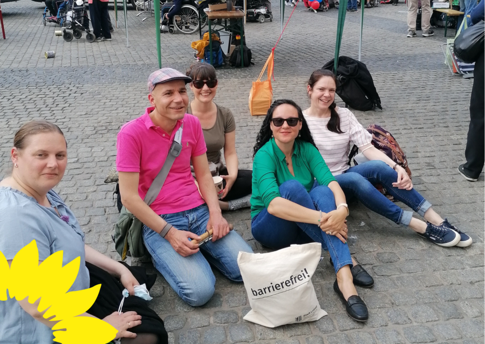 !B 6 bFrauen und Männer sitzen auf dem Marktplatz vor ihnen steht ein Beutel mit der Aufschrift "Barrierefreiheit"