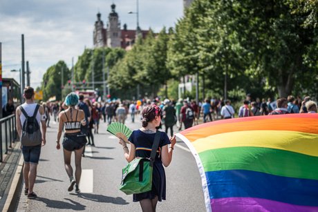 Auf dem Foto ist eine Menschenmenge zu sehen (von hinten) welche vor dem unscharfen Hintergrund, dem Leipziger neuen Rathaus, mit Regenbogenfahnen und bunten Luftballons demonstriert.