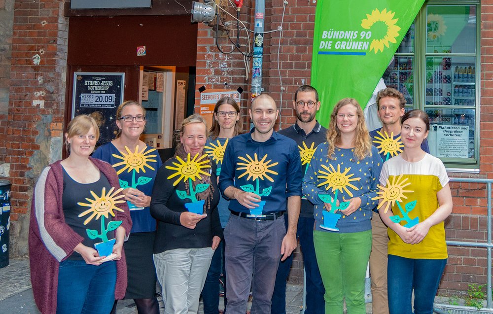 Auf dem Bild stehen neun Personen mit einer Holzsonnenblume in der Hand vor dem Eingang der Halle D des Werk 2 in Leipzig.