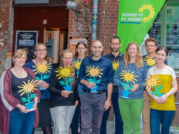 Auf dem Bild stehen neun Personen mit einer Holzsonnenblume in der Hand vor dem Eingang der Halle D des Werk 2 in Leipzig.