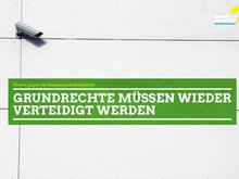 Auf dem Foto ist eine Häuserwand zu sehen mit einer angebrachten Überwachungskamera. Mittig ist ein grüner Kasten zu sehen in welchem folgender Schriftzug steht: "Grundrechte müssen wieder verteidigt werden."